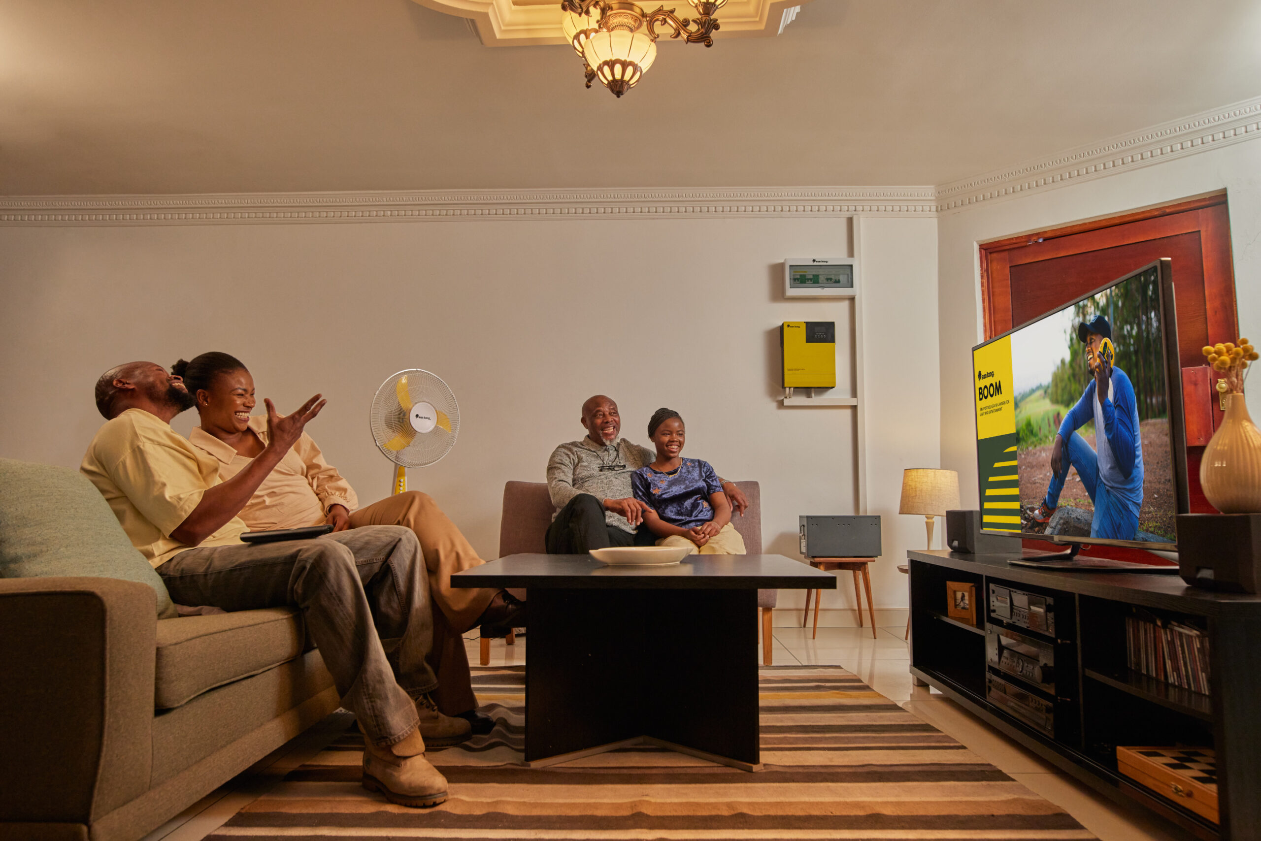 Family in a living room enjoying their solar powered Sun King rooftop inverter system, which is powering appliances like a fan and TV.