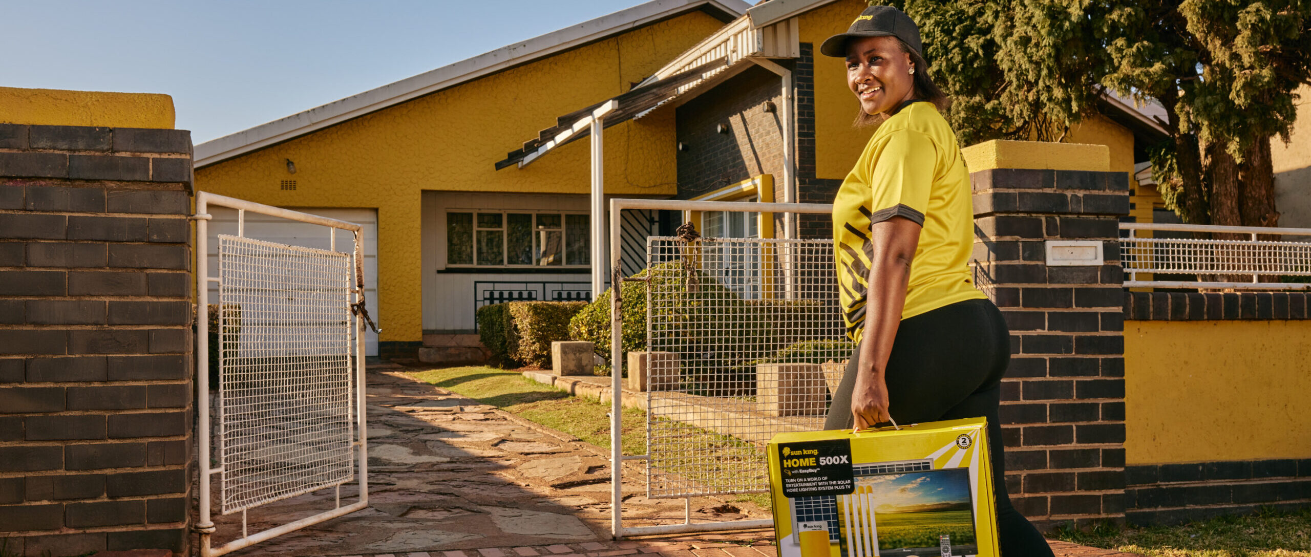 Sun King employee walks through a gate to a customer's house, carrying a Sun King Home 500X home solar powered lighting system complete with solar panels.