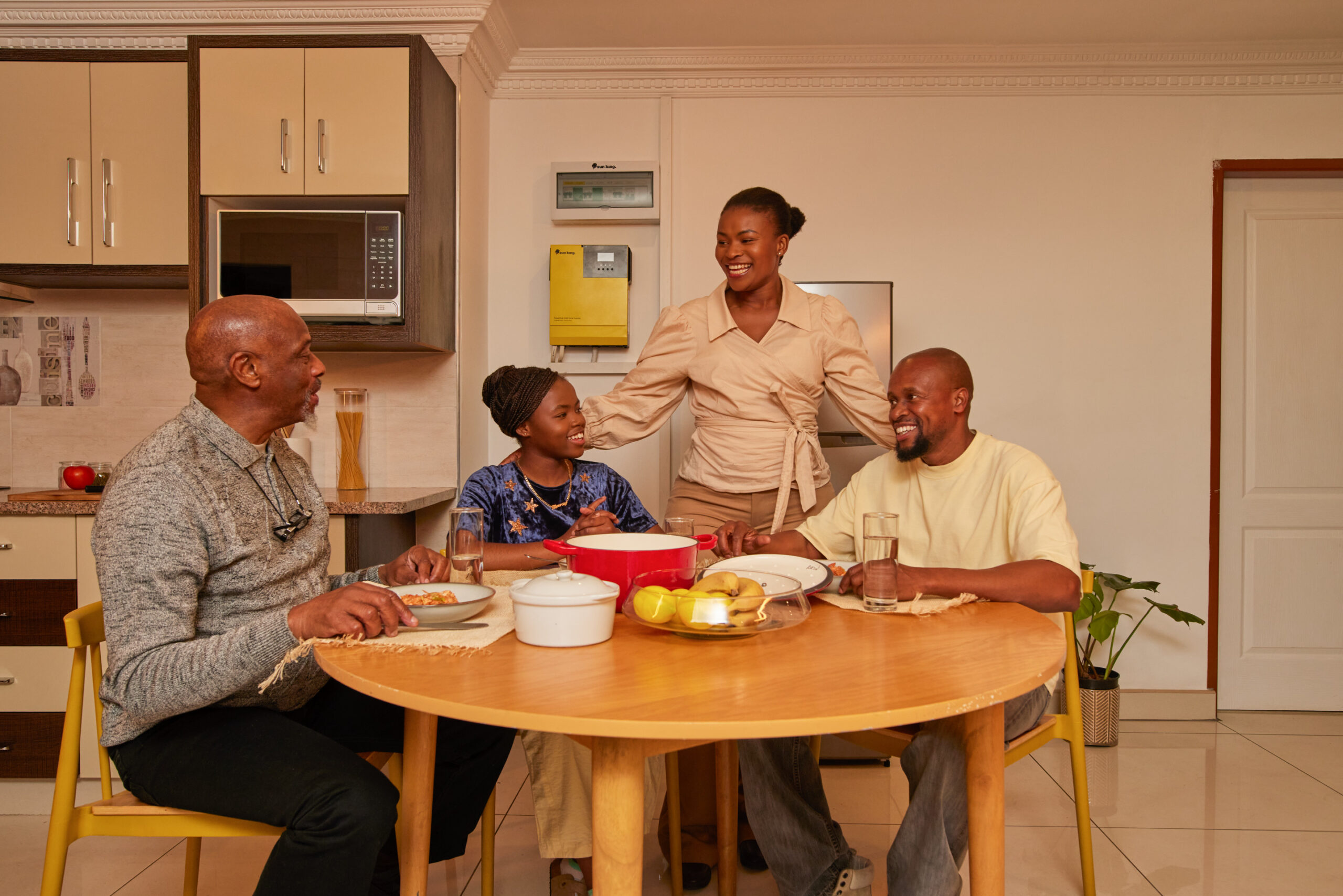 A family sits in their well-lit living room, which features a fridge, microwave and other appliances powered by a Sun King solar inverter home system.